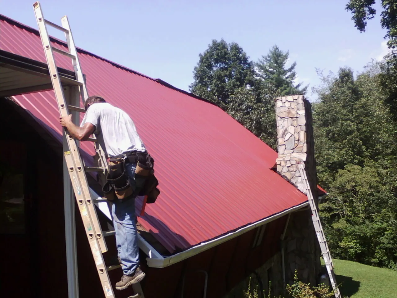 Roofer on a ladder beside a red metal roof and stone chimney — Soffit & Fascia Repair by Roofing Brothers in Oberlin